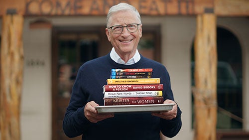 Bill Gates standing with a stack of books on a tray in front of a 